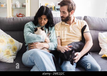 Content young couple in casual outfits resting on sofa together and caressing cute cats while spending time at home during weekend Stock Photo