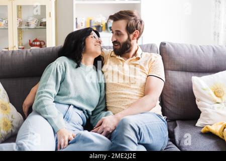 Cheerful young couple in casual outfits smiling and looking at each other while cuddling on sofa together Stock Photo