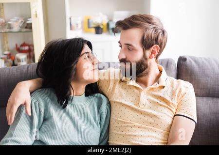 Cheerful young couple in casual outfits smiling and looking at each other while cuddling on sofa together Stock Photo