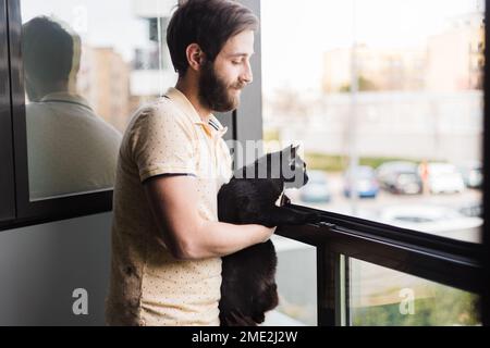 Side view of calm young bearded male owner in casual t shirt with black cat standing near window while admiring cityscape in daytime Stock Photo