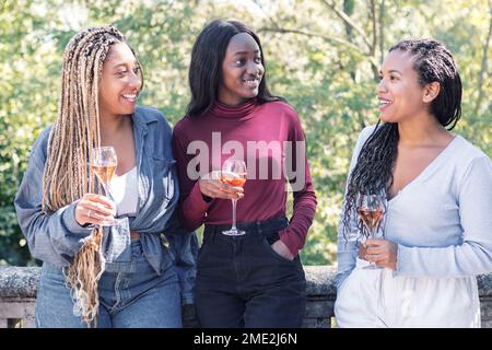 Happy multiracial female friends in casual clothes standing on terrace with glasses of champagne on sunny day in park Stock Photo