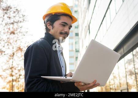 From below side view of positive young Hispanic male engineer in smart casual clothes and yellow helmet working on laptop while standing near modern b Stock Photo
