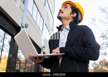 From below side view of positive young Hispanic male engineer in smart casual clothes and yellow helmet working on laptop while standing looking up ne Stock Photo