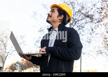 From below side view of positive young Hispanic male engineer in smart casual clothes and yellow helmet working on laptop while standing looking up ne Stock Photo