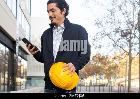 Crop young confident ethnic male engineer in smart casual wear with dark hair and beard smiling and using smartphone while standing on city street wit Stock Photo
