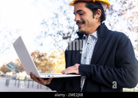 From below side view of positive young Hispanic male engineer in smart casual clothes and yellow helmet working on laptop while standing near modern b Stock Photo