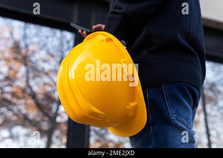Side view of crop unrecognizable male engineer in casual clothes with yellow helmet in hand standing near modern building and messaging on smartphone Stock Photo