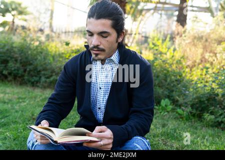 Focused young ethnic male with dark hair and beard in smart casual wear sitting on grassy lawn and reading interesting book in park Stock Photo