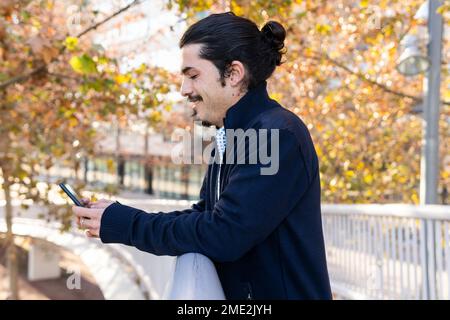 Side view of delighted young Hispanic male with dark hair and beard in smart casual outfit smiling while standing on footbridge and messaging on smart Stock Photo