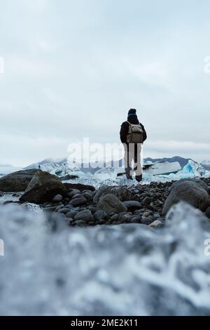 Ground level of unrecognizable tourist in outerwear with backpack holding hands in pockets while standing on stony coast covered with ice against over Stock Photo