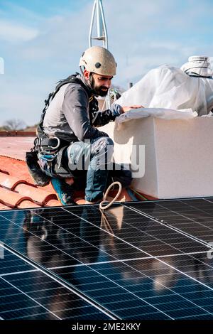 Adult bearded mechanic in hat sitting on tiled roof with shiny just installed solar battery in residential city area Stock Photo
