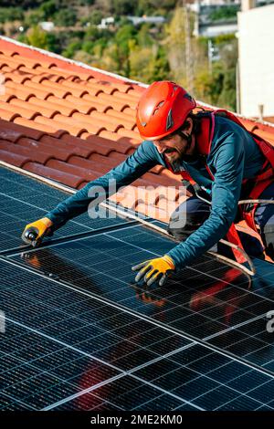 Male worker in protective gloves and helmet on rooftop of house screwing solar panels during installation on rooftop Stock Photo