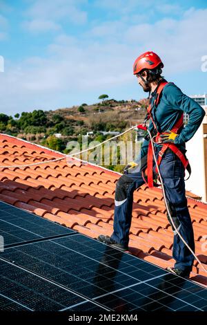 Full body of male worker in overalls and helmet standing with wire in hand while installing solar panels on tiled roof of house in sunlight Stock Photo