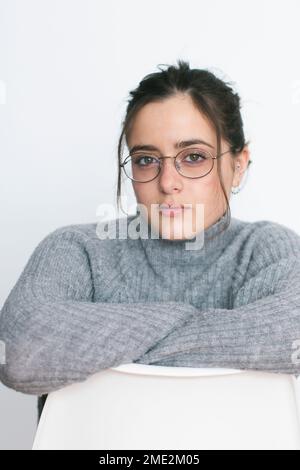 Female with dark hair in warm gray sweater and glasses standing near white wall looking at camera Stock Photo