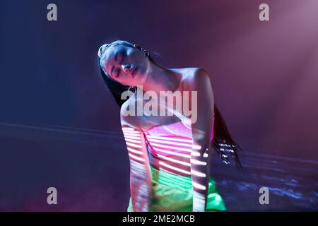 Young female in pink top and light green pants with eyes closed in studio with blue neon illumination and smoke Stock Photo