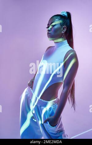Young female model in white bodysuit standing with one hand on waist and another in pocket against violet background with light lines from projector Stock Photo