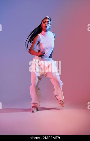 Full body of active female in white clothes dancing in studio against purple neon illumination looking at camera Stock Photo