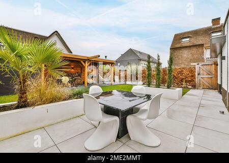 Black marble table with white chairs located on tiled floor near planter with palm tree on cloudy autumn day in daytime Stock Photo