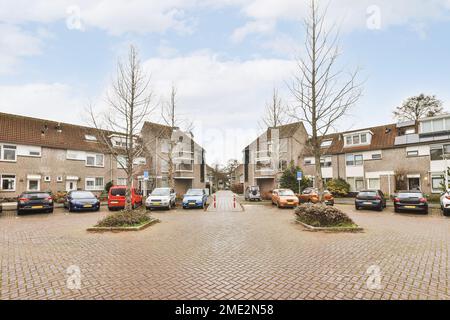 Cars parked on asphalt parking lot near paved road trees and brick buildings Stock Photo