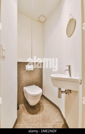 Interior of bathroom with white walls and brown tiled floor with ceramic toilet and small sink Stock Photo