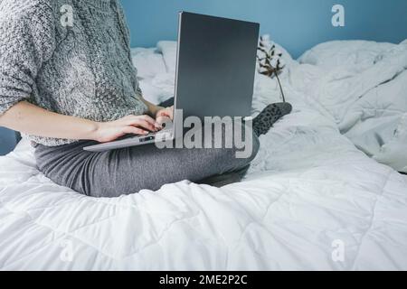 Cropped unrecognizable woman sitting on bed and typing on laptop keyboard while working on remote project at home Stock Photo