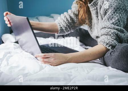 Cropped unrecognizable woman sitting on bed and typing on laptop keyboard while working on remote project at home Stock Photo