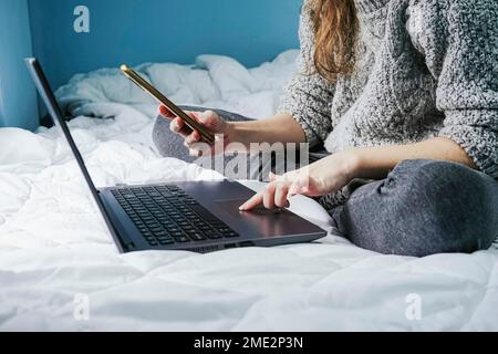 Side view of anonymous female freelancer sitting on bed and using mobile phone while working on netbook Stock Photo