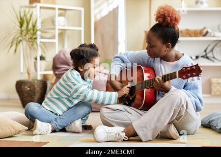Full length side view portrait of caring mother playing guitar with daughter at home and teaching little girl Stock Photo