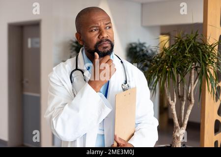 Thoughtful african american male doctor with clipboard in hospital corridor Stock Photo