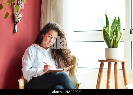 Woman relaxing in a armchair writing a diary indoors surrounded by plants Stock Photo