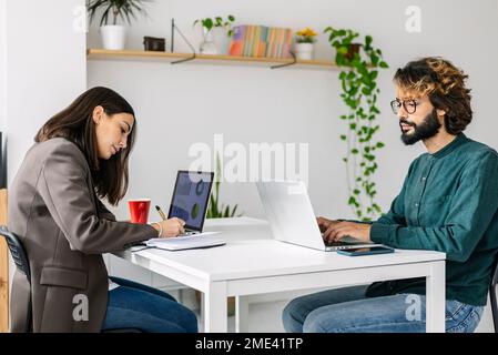 Businessman using laptop by colleague writing in diary at desk Stock Photo