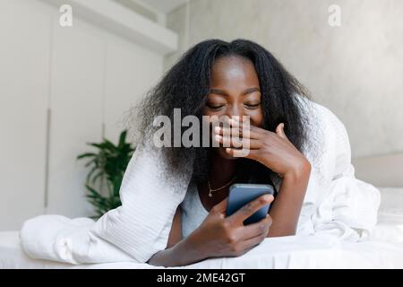 Happy young woman looking at mobile phone lying on bed Stock Photo