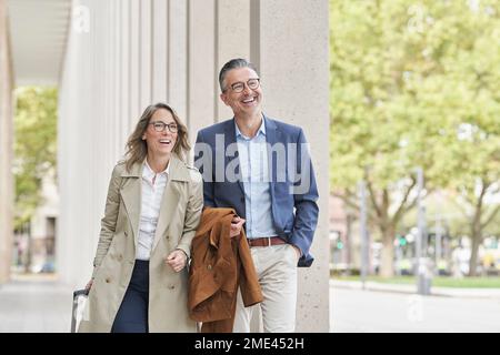 Happy mature businesswoman with suitcase walking with colleague Stock Photo