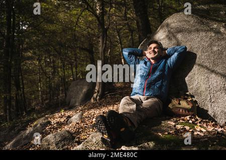 Happy mature man with hands behind head relaxing by rock in forest Stock Photo