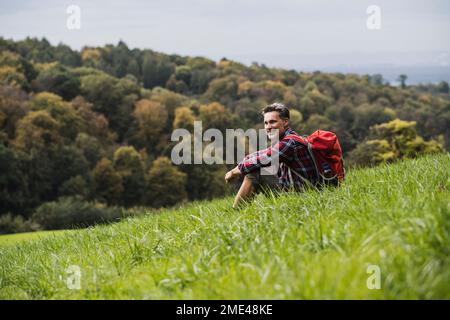 Smiling man with backpack sitting on grass Stock Photo