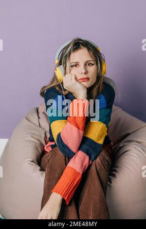 Sad girl listening to music through headphones sitting on bean bag against purple background Stock Photo