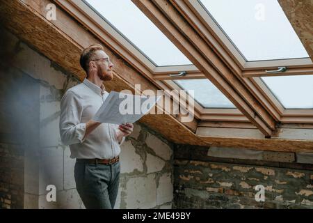 Thoughtful architect standing with blueprint looking through window at construction site Stock Photo