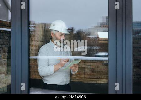 Businessman wearing hardhat making notes at construction site seen through glass Stock Photo