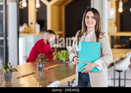 Smiling businesswoman with file folder standing by desk in office Stock Photo