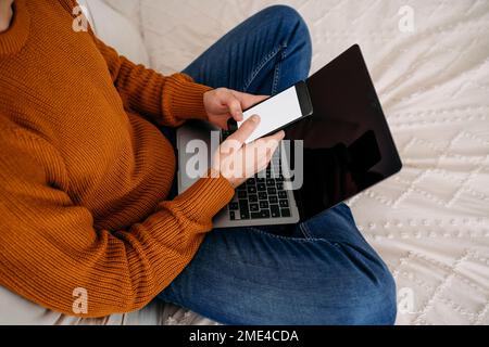 Hands of freelancer using smart phone sitting with laptop on bed at home Stock Photo