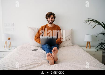 Happy young freelancer sitting on bed with laptop Stock Photo