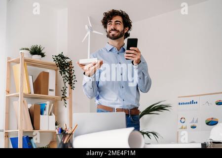 Thoughtful engineer with wind turbine and mobile phone standing in office Stock Photo