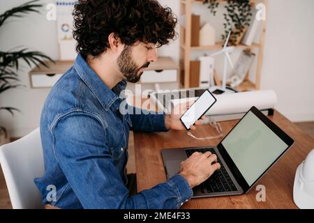 Engineer working on smart phone and laptop sitting at desk in office Stock Photo
