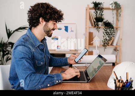Smiling engineer working on laptop and smart phone sitting at desk in office Stock Photo