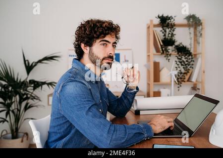 Contemplative engineer holding eyeglasses with laptop on desk in office Stock Photo