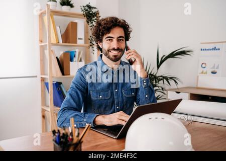 Happy engineer talking on mobile phone with laptop at desk in office Stock Photo
