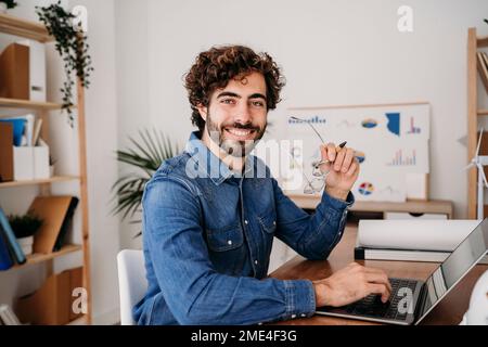 Happy young engineer holding eyeglasses with laptop on desk in office Stock Photo