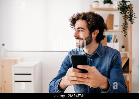 Thoughtful smiling young engineer holding smart phone sitting at desk in office Stock Photo