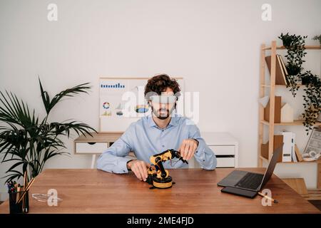 Young engineer wearing VR glasses holding robotic arm on desk in office Stock Photo