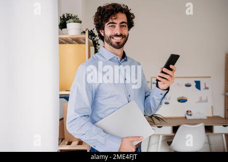 Smiling young engineer holding smartphone and laptop in office Stock Photo